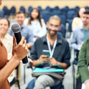 istockphoto-1487907475-612×612 Cropped shot of a businesswoman holding a mic speaking to group of audience during a business conference